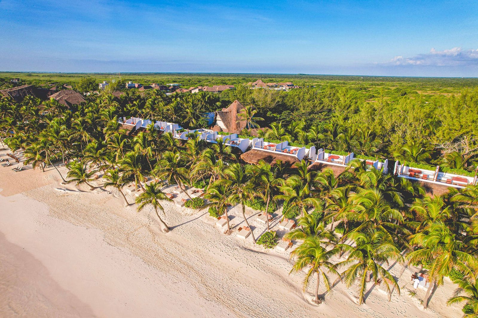 A high-angle drone view of The Beach Tulum Hotel, showcasing beachfront rooms with private rooftop terraces. Each terrace features a hot tub, sofa, and loungers, all facing the beautiful blue Tulum beach. Lush jungle foliage forms the backdrop behind the hotel.