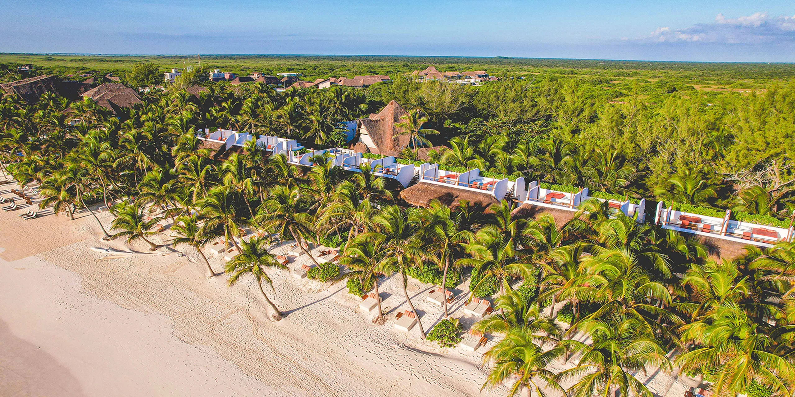 A high-angle drone view of The Beach Tulum Hotel, showcasing beachfront rooms with private rooftop terraces. Each terrace features a hot tub, sofa, and loungers, all facing the beautiful blue Tulum beach. Lush jungle foliage forms the backdrop behind the hotel.